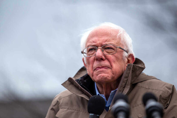 shutterstock_16979420741.jpg featured image Bernie Sanders wearing glasses speaking outdoors with microphones.