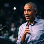 Barack Obama in blue shirt speaking at podium with microphone.