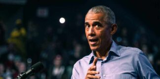 Barack Obama in blue shirt speaking at podium with microphone.
