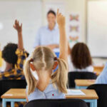 Children raising hands in classroom, teacher in background
