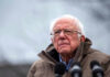 Bernie Sanders wearing glasses speaking outdoors with microphones.