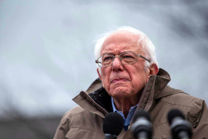 Bernie Sanders wearing glasses speaking outdoors with microphones.