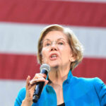 A female politician speaking passionately into a microphone against a backdrop of an American flag