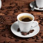 A white coffee cup filled with coffee on a wooden table surrounded by coffee beans