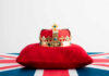 A royal crown resting on a red velvet pillow with a British flag background