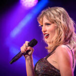 Taylor Swift performing with microphone on stage, colorful lighting.