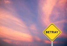 A yellow warning sign with the word 'BETRAY' against a colorful sunset sky