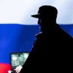 Silhouette of a military personnel in front of the Russian flag using a laptop