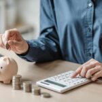 Person placing a coin into a piggy bank while using a calculator