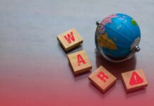 Iran War CHOKES Lifeline – Millions Could Starve A small globe next to wooden blocks spelling 'WAR' with a warning symbol