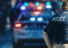 Police officer stands near patrol car with lights flashing.