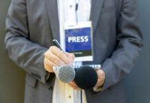 Journalist with press badge, notebook, and microphones.