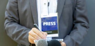 Journalist with press badge, notebook, and microphones.