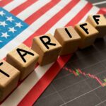 Wooden blocks spelling 'TARIFFS' on a background of the US flag and financial charts