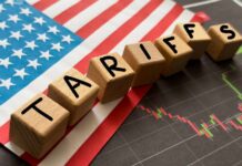 Wooden blocks spelling 'TARIFFS' on a background of the US flag and financial charts
