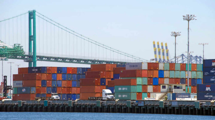 A view of stacked shipping containers at a port with a bridge in the background