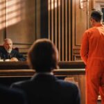 Defendant in orange jumpsuit stands before a judge in a courtroom
