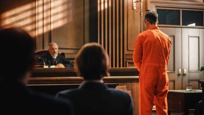 Defendant in orange jumpsuit stands before a judge in a courtroom
