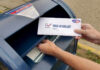 Hands mailing a ballot envelope into a blue mailbox