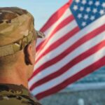 Pentagon Orders THESE Tests For Troops Over 30 Soldier in uniform standing in front of an American flag by the ocean