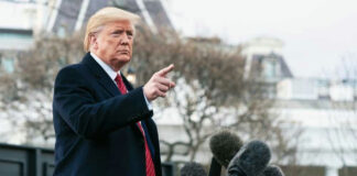 Man speaking to microphones, pointing outdoors.