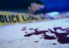Blood on pavement with police tape and a hand in the background