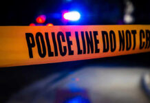 Horror at Youth Game – Case Stalls Police tape with flashing lights in the background.